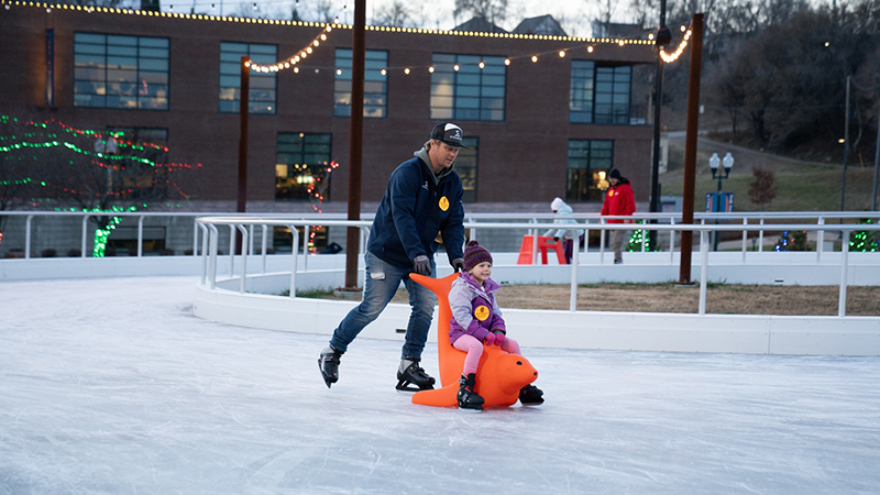 A father ice skating with daughter using a seal skate helper